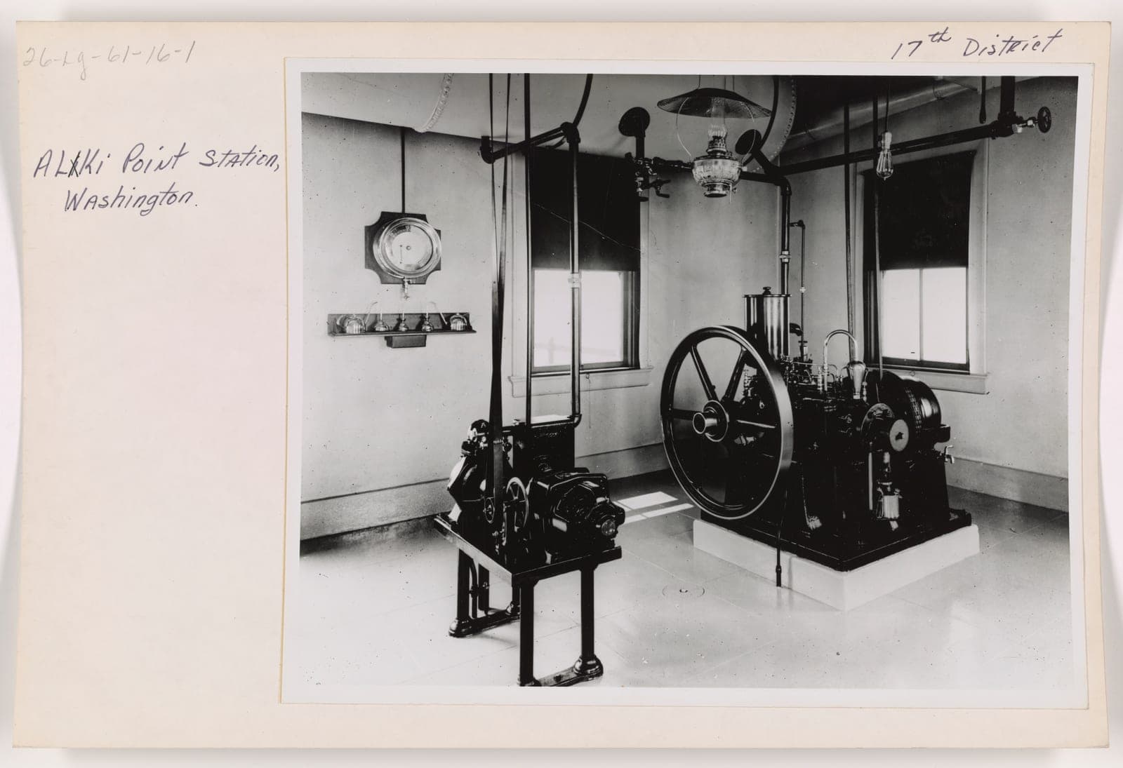 A black and white photograph, annotated by hand "Alki Point Station, Washington. 17th District", of a tiled equipment room containing a large flywheel engine on a plinth and a smaller machine on a steel frame, with a gauge on the wall and an oil lamp hanging from the ceiling.